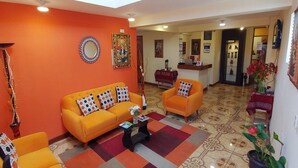 A brightly colored living area with orange sofas and patterned cushions, a tiled floor, and a reception desk in the background. - Qorikilla Hostal (Cusco)