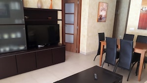 A modern living room with a flat-screen TV on a dark wooden cabinet, a black coffee table, and a dining area with a wooden table and black chairs. - Oropesa Ciudad de Vacaciones 3000 (Oropesa del Mar)