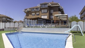 A swimming pool with a deck, a covered seating area, and a building with balconies in the background. - Gala (Noja)