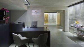 Un área de recepción moderna con un mostrador, dos sillas grises, un letrero que dice "NH COLLECTION PALOMINE", y un pasillo que lleva a una sala con una mesa de vidrio que exhibe bebidas y aperitivos.