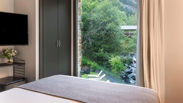 A bedroom with a bed, a TV, a wardrobe, and a window with a view of greenery and a building. - NH Collection Andorra Palome (Arinsal)