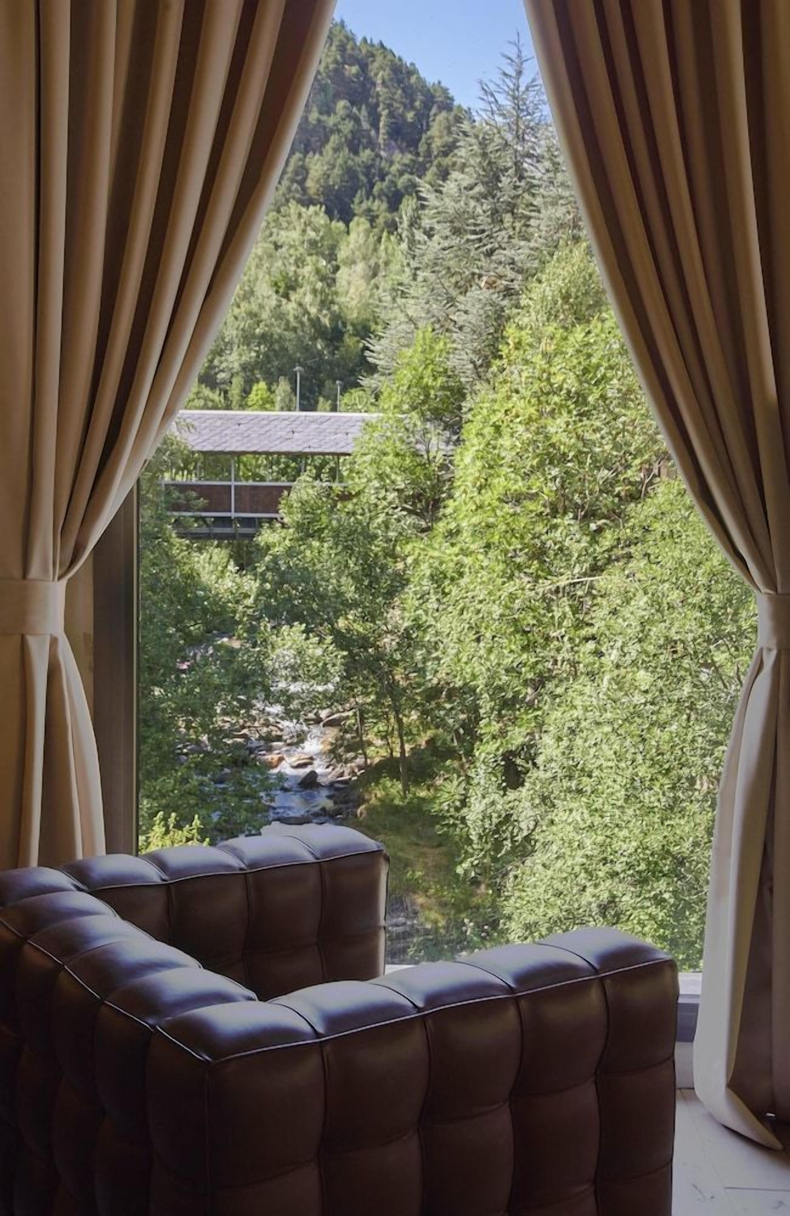Una habitación con vistas a una ladera boscosa, una cascada pequeña y un edificio a lo lejos. Hay dos sillones tapizados en primer plano.