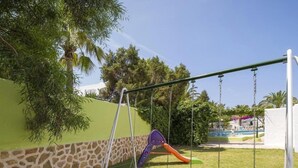 A playground with swings and a slide, surrounded by a green wall and trees. - Hotel Bahia Playa (Sant Josep de sa Talaia)