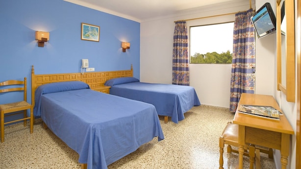 A room with two beds, a wooden desk, a chair, a window with curtains, and a wall-mounted TV. - Hotel Bahia Playa (Sant Josep de sa Talaia)