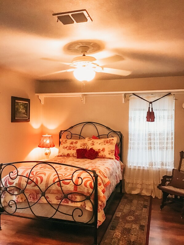 Un dormitorio con cama, ventilador de techo, mesita de noche con lámpara, ventana con cortinas y una silla.