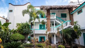 A two-story building with white walls and teal accents, surrounded by lush tropical vegetation. - Hotel Costa 55 (Puerto Escondido)