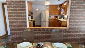 A dining area with a brick wall, wooden table, and chairs. A kitchen is visible through a mirror above the table.