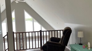 Ein Zimmer mit hoher Decke, ein dunkles Sessel, ein türkisner Nachttisch mit Lampe und Blick auf einen Balkon mit Geländer.