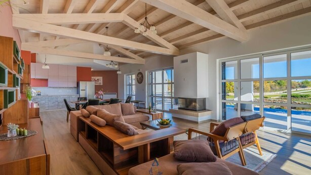 A spacious living area with a wooden ceiling, a fireplace, and large windows offering a view of the outdoors. - Modern 5-BR villa for 10+1 with heated pool and specious land (Filipana)