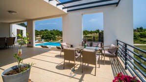 A balcony with a pool, wicker furniture, and a view of greenery. - Modern 5-BR villa for 10+1 with heated pool and specious land (Filipana)