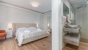 Habitación de hotel moderna con una cama grande, una mesita de noche, un tocador, un baño con lavabo y espejo, y una ducha.