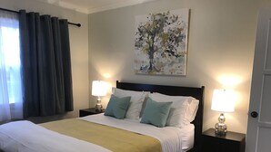 Un letto rifatto con cuscini bianchi e verdi, due abat-jour e un quadro raffigurante un albero al muro.
