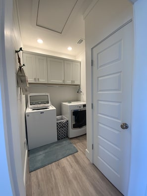 Compact laundry room with a washing machine, dryer, and trash bin, featuring cabinetry and a wooden floor.
