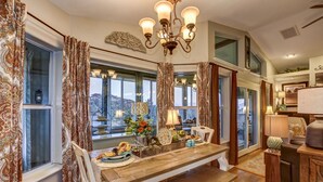 A dining area with a wooden table, chairs, and a chandelier. There are windows with curtains, a view of the outside, and a small side table with a lamp. - The Retreat at Thumb Butte (Prescott)