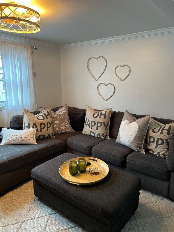 Un salon moderne avec un canapé d’angle agrémenté de coussins ornés de la mention « OH HAPPY DAY », une table basse en bois décorée d’objets d’art et des décorations murales en forme de cœur.