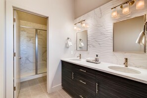 Une salle de bain moderne avec deux vasques, des meubles de rangement aux couleurs sombres, un grand miroir et une cabine de douche en verre.