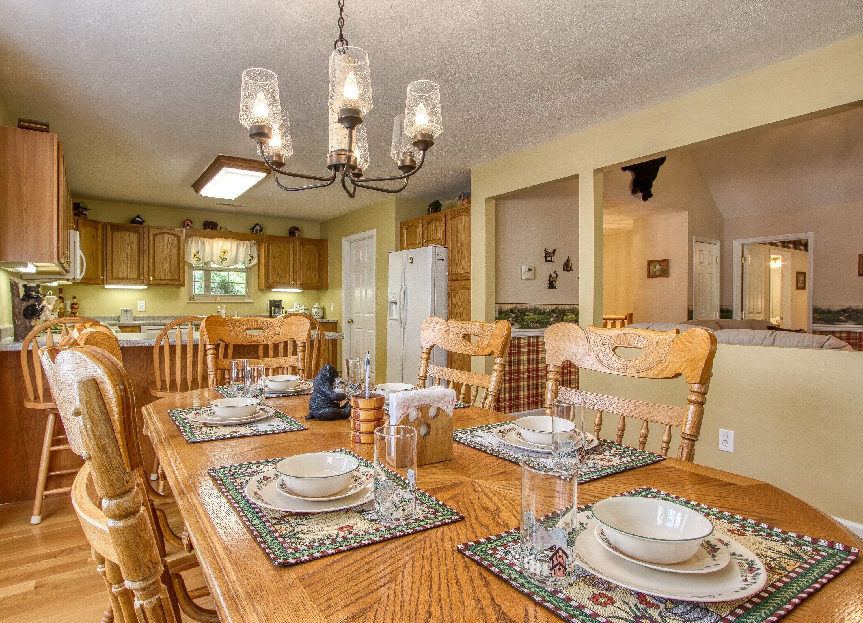 A dining area with a wooden table set for four, surrounded by wooden chairs. A chandelier hangs above, and the room has wooden cabinets and a kitchen in the background.
