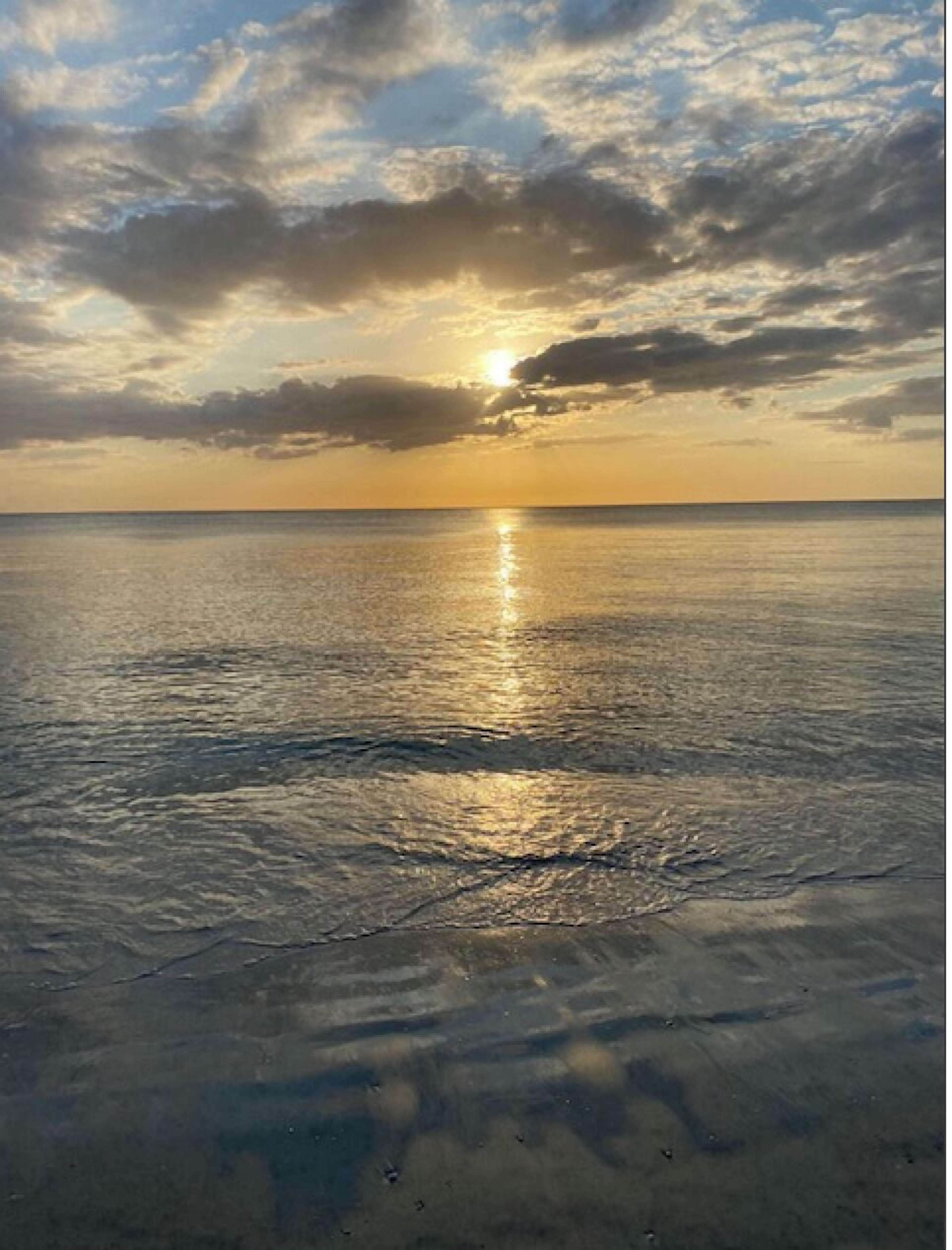 A beach with a calm sea reflecting the sun, and a clear sky with clouds.