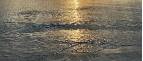A beach with a calm sea reflecting the sun, and a clear sky with clouds.
