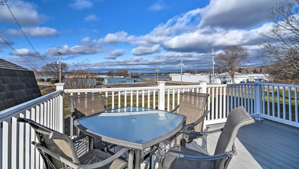 A deck with a glass table and chairs, white railings, and a view of a body of water and buildings in the distance. - Downtown Boyne City Home, Walk to Lake Charlevoix! (Boyne City)