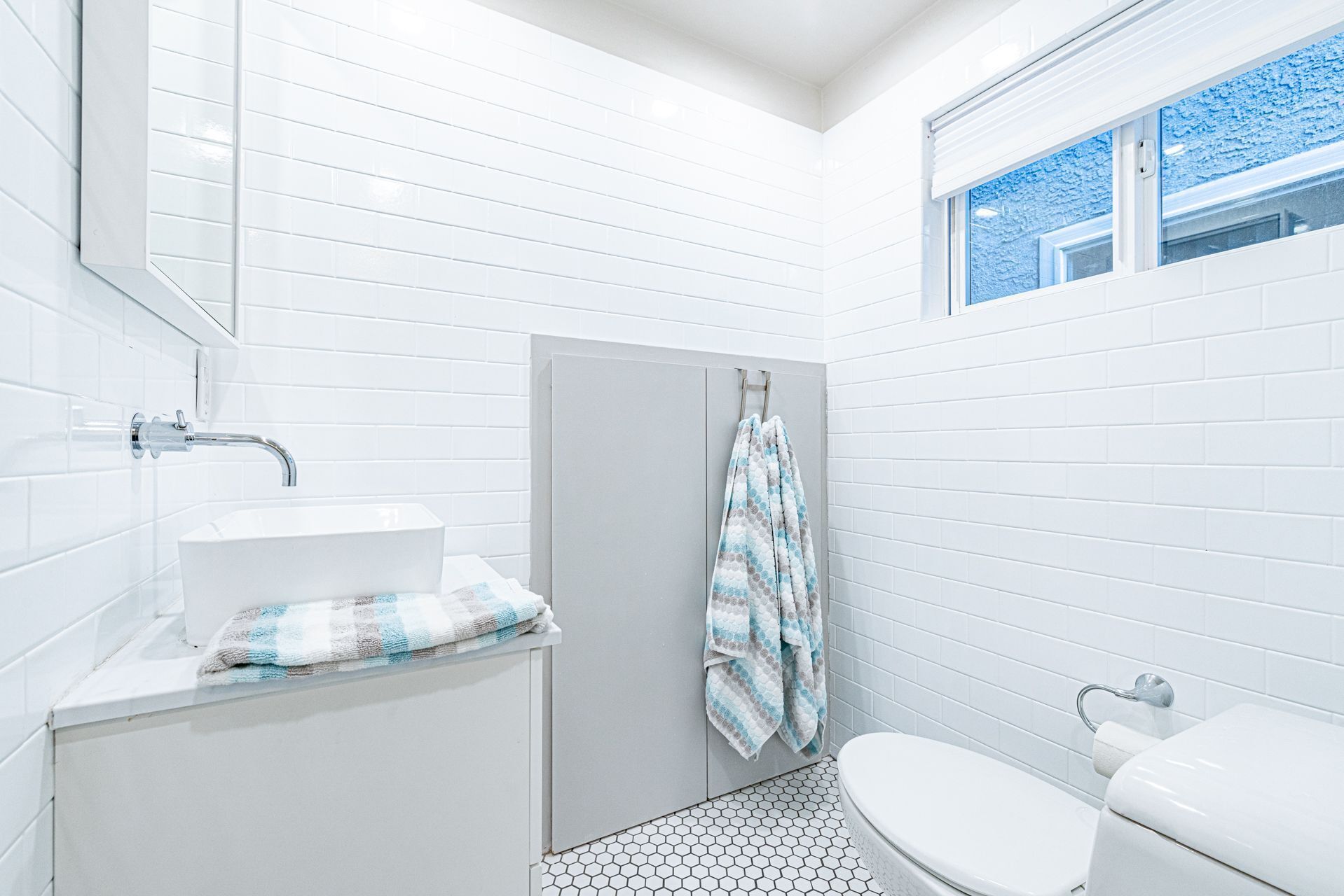 A small, modern bathroom with white tiles, a white sink, a toilet, and a shower area.
