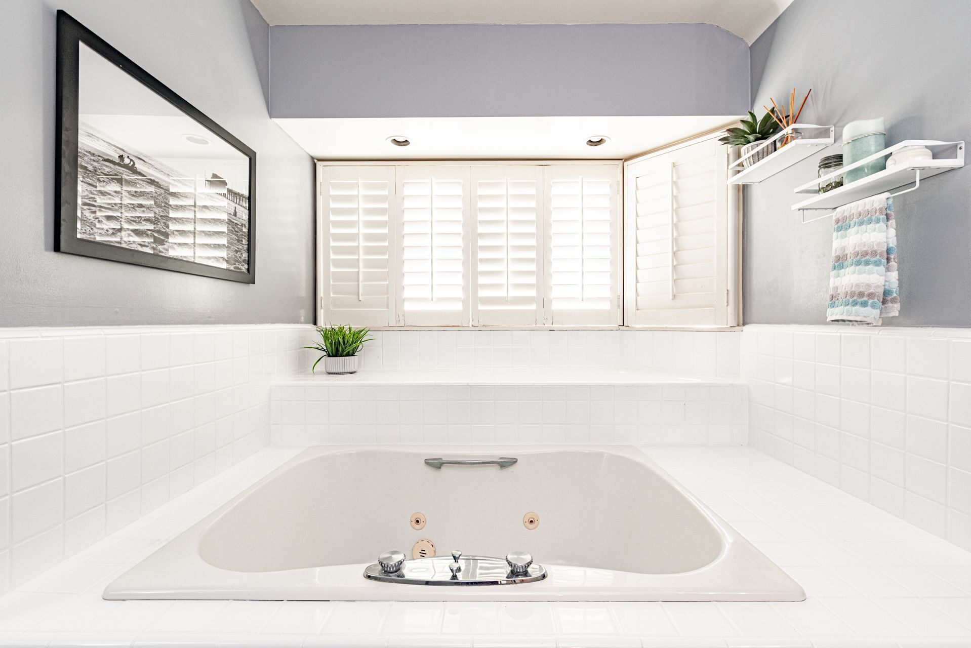 A modern bathroom with a large bathtub, white tiled walls, a framed picture on the wall, a window with shutters, and a shelf with toiletries.