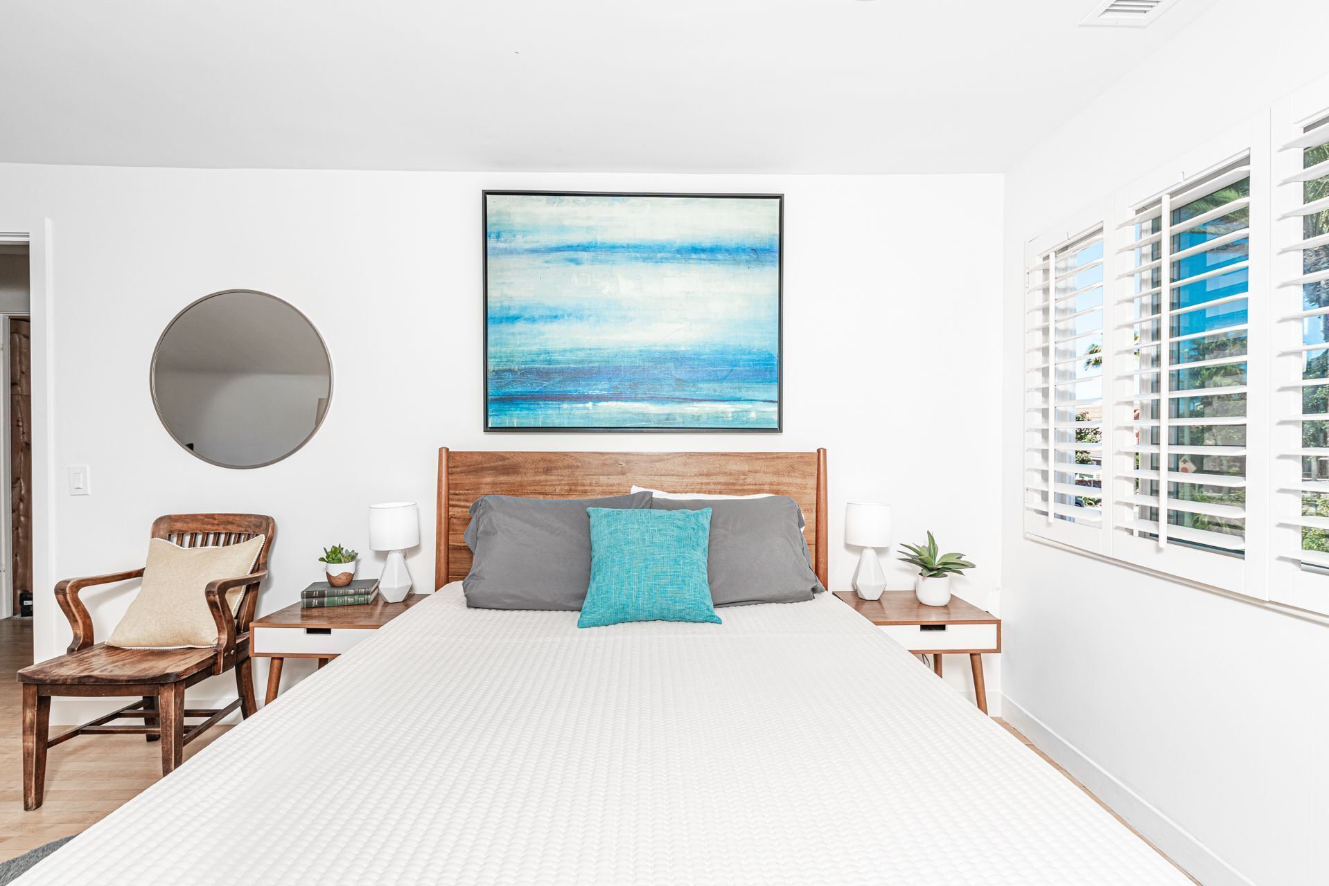 A bedroom with a large bed, a wooden headboard, a round mirror, a chair, a bedside table with a lamp, a small plant, and a window with blinds.