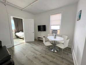 A hotel room with a bed, a TV, a dining table with chairs, a small table with a plant, and a window with blinds. - Modern Beachfront Suite 306 (Ocean City)