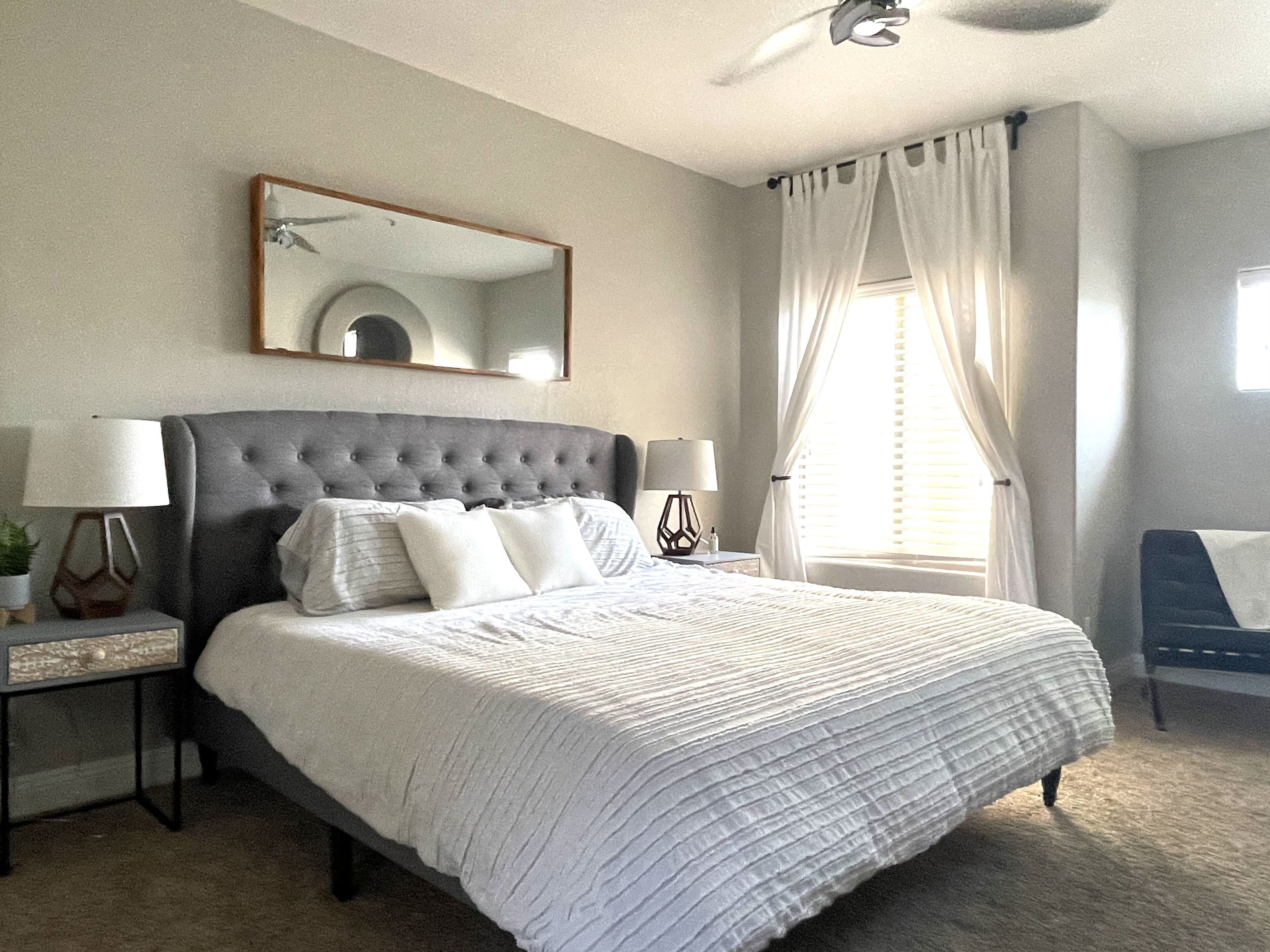 Une chambre à coucher bien rangée, avec un grand lit, deux tables de chevet, un miroir et une fenêtre avec des rideaux.