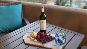 Een tafel met een fles wijn, kaas, aardbeien en een glas water op een balkon met uitzicht op een zwembad en groen.