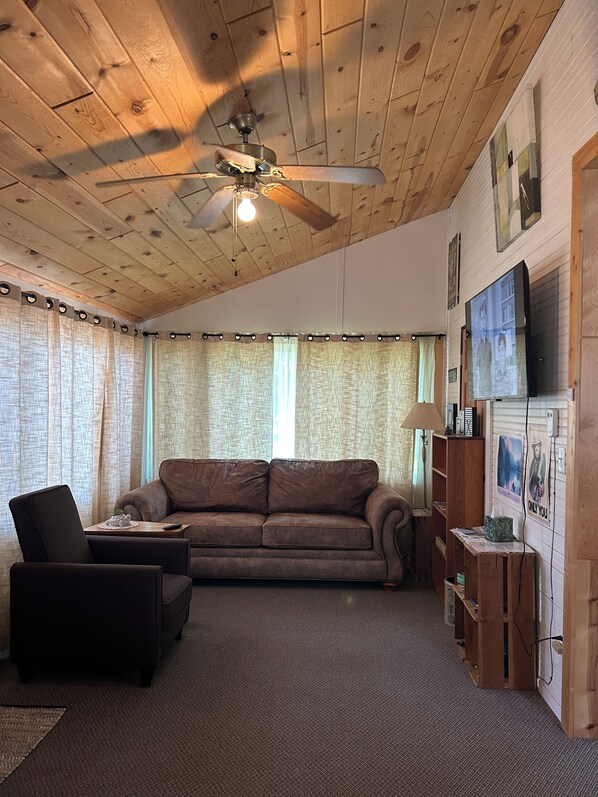 A cozy living room with a wooden ceiling, a ceiling fan, a brown leather sofa, a black armchair, and a wooden sideboard with a television.