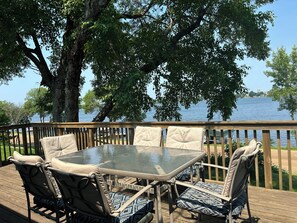 A deck with a glass-top table and cushioned chairs, overlooking a body of water.