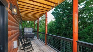 Un patio in legno con vasca idromassaggio, barbecue e ringhiera metallica che si affaccia su una zona boschiva.