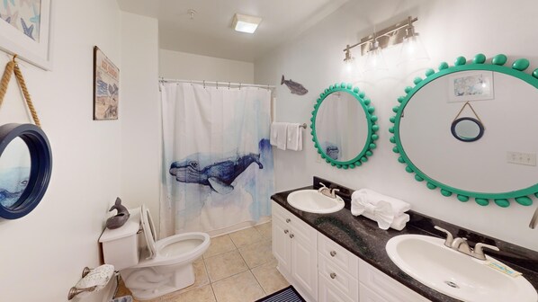 Une salle de bain avec deux lavabos, des toilettes, un rideau de douche orné de motifs de baleines et plusieurs miroirs avec un cadre vert.