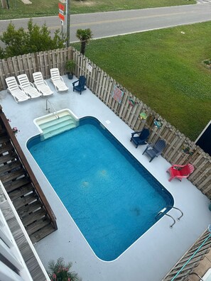 A rectangular swimming pool with a wooden deck, surrounded by lounge chairs and a wooden fence.
