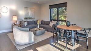 A modern living room with a grey sofa, a round wooden table, and a large window with a view of trees. - Luxury Condo, Downtown Asheville (Asheville)