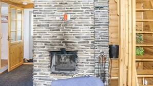 Una chimenea de piedra con res protector metálico, vigas de madera y un sillón azul en una habitación con paredes de madera y una puerta.