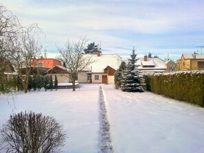 A residential street with houses, a snow-covered path, and a hedge.