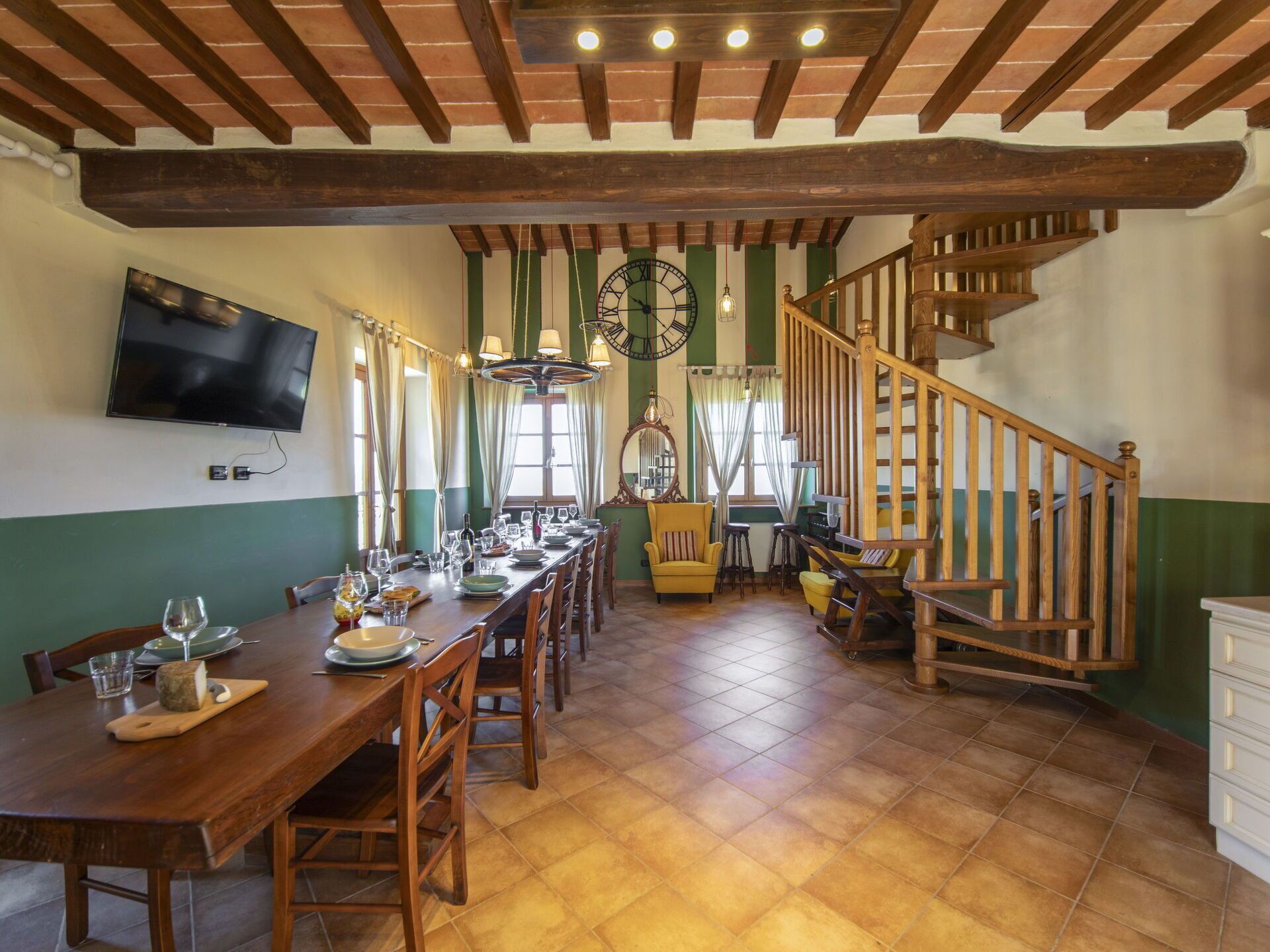 A dining area with a long wooden table set for a meal, a clock on the wall, and a wooden staircase.