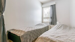 Ein kleiner, schlichter Raum mit zwei Einzelbetten, einem Fenster mit Vorhängen und einem Heizkörper.