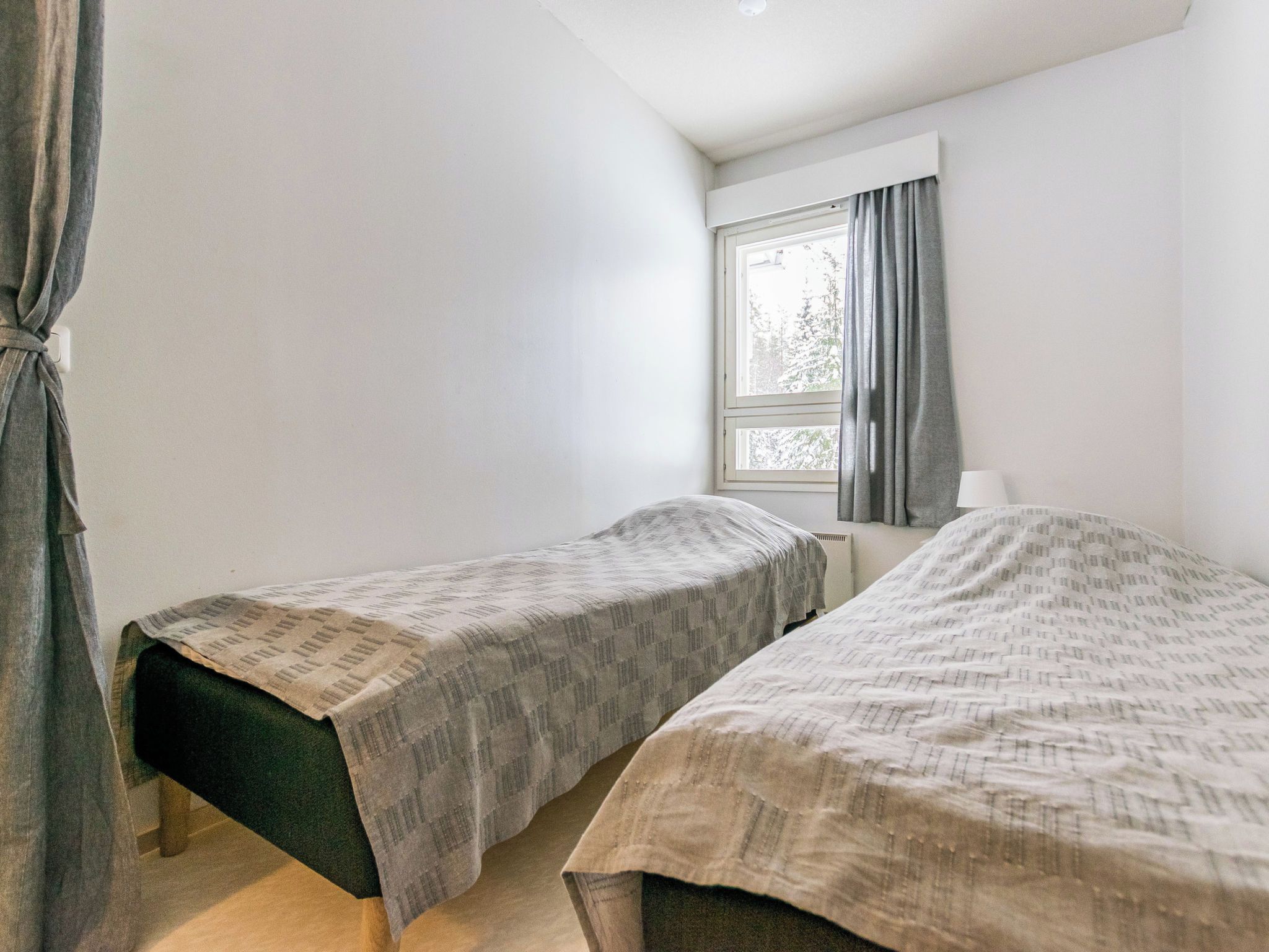 A small, simple room with two single beds, a window with curtains, and a radiator.