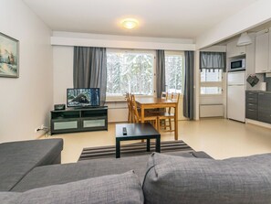 Un salon moderne avec un canapé, un téléviseur posé sur un meuble, un coin repas avec des chaises, une cuisine équipée et une grande fenêtre avec des rideaux.