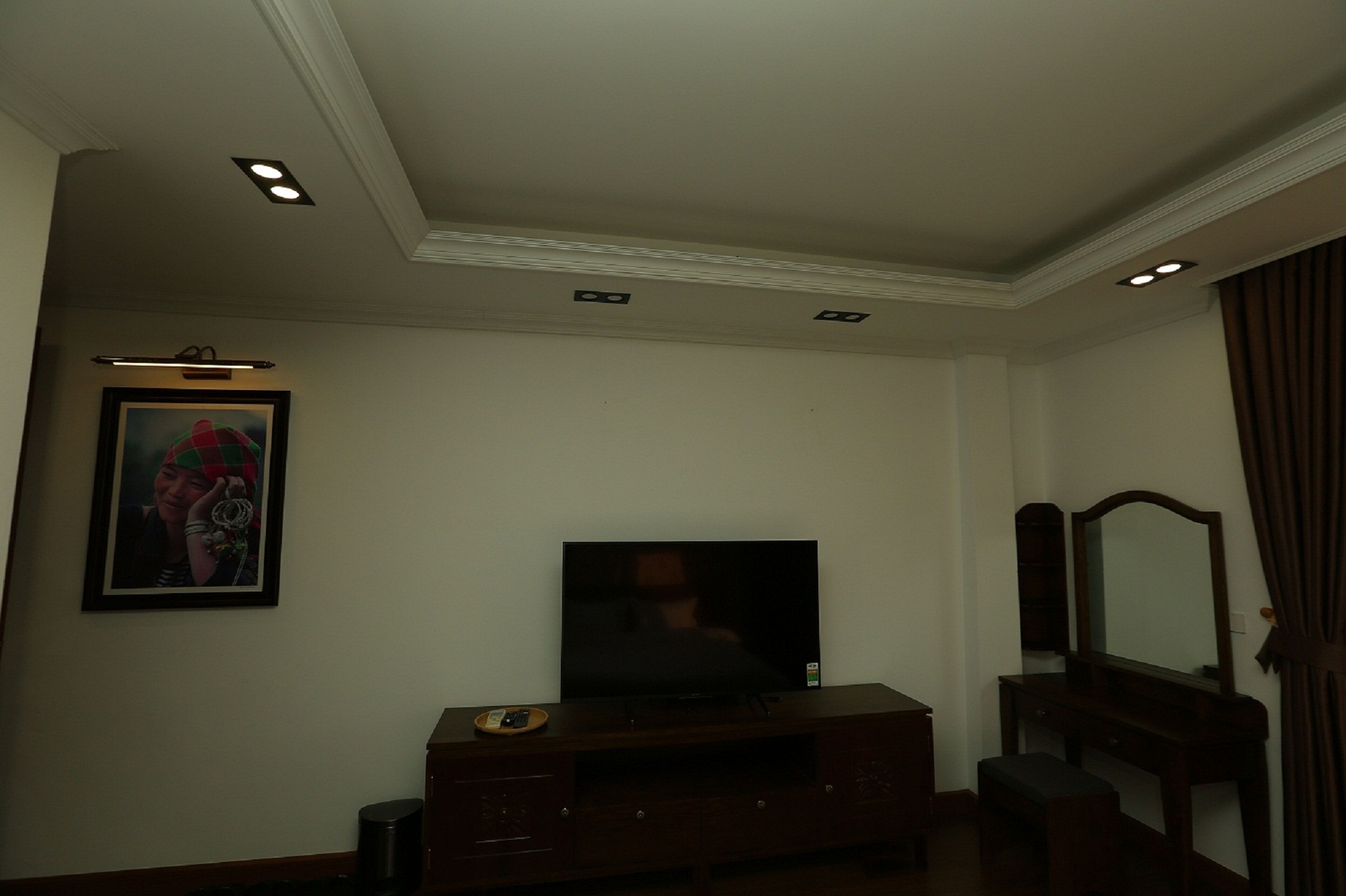 A hotel room with a flat-screen TV on a wooden stand, a framed picture on the wall, a mirror, and a small bench.