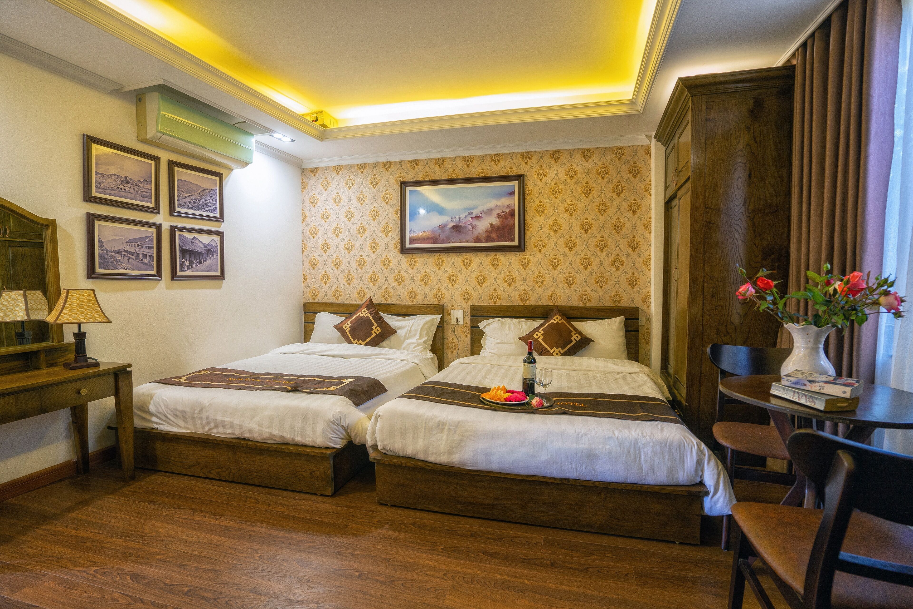 A hotel room with two beds, a desk, a chair, a vase with flowers, and a painting on the wall.