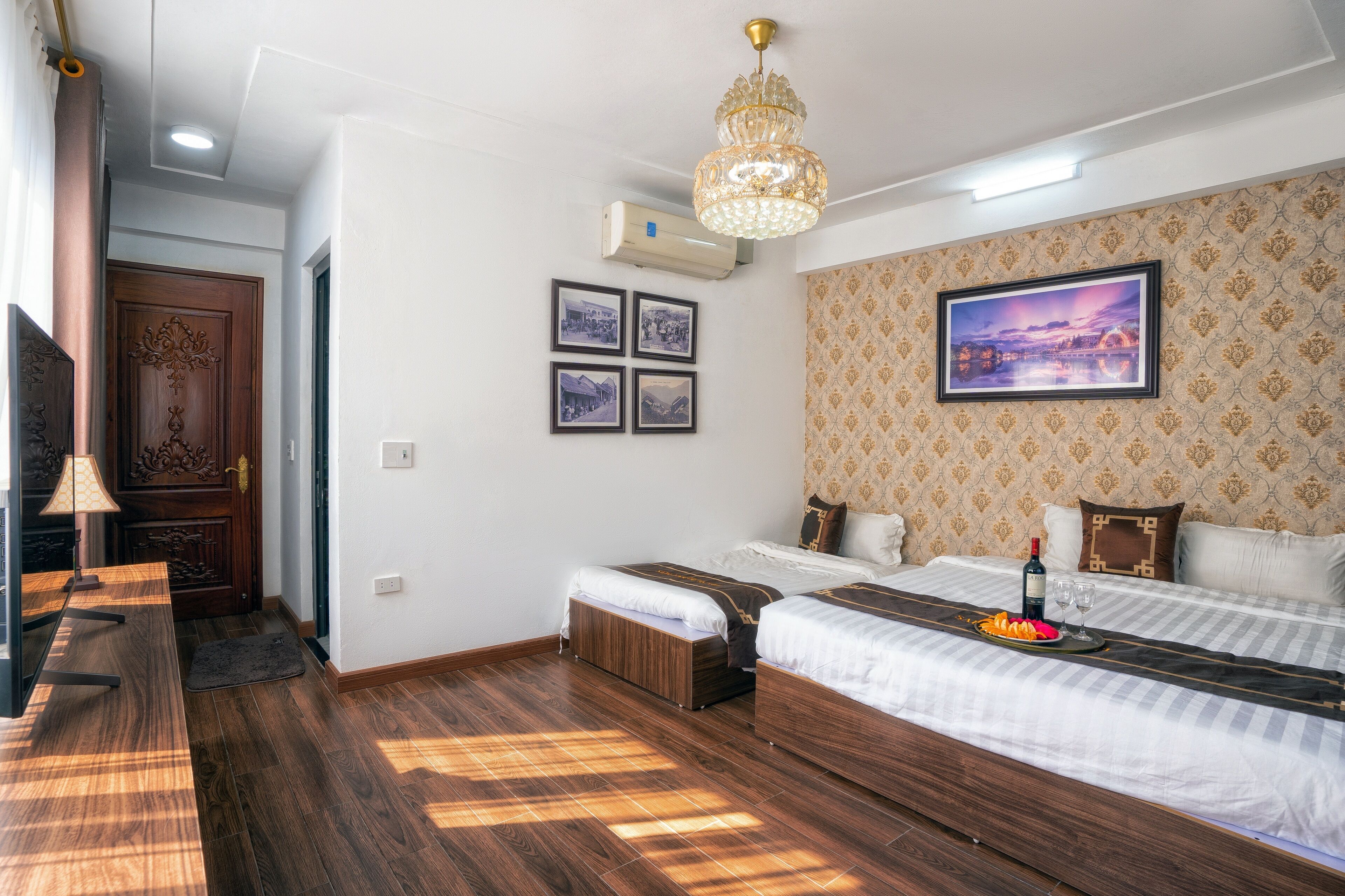 A hotel room with two beds, a wooden floor, a chandelier, a wall-mounted air conditioner, and a framed picture on the wall.