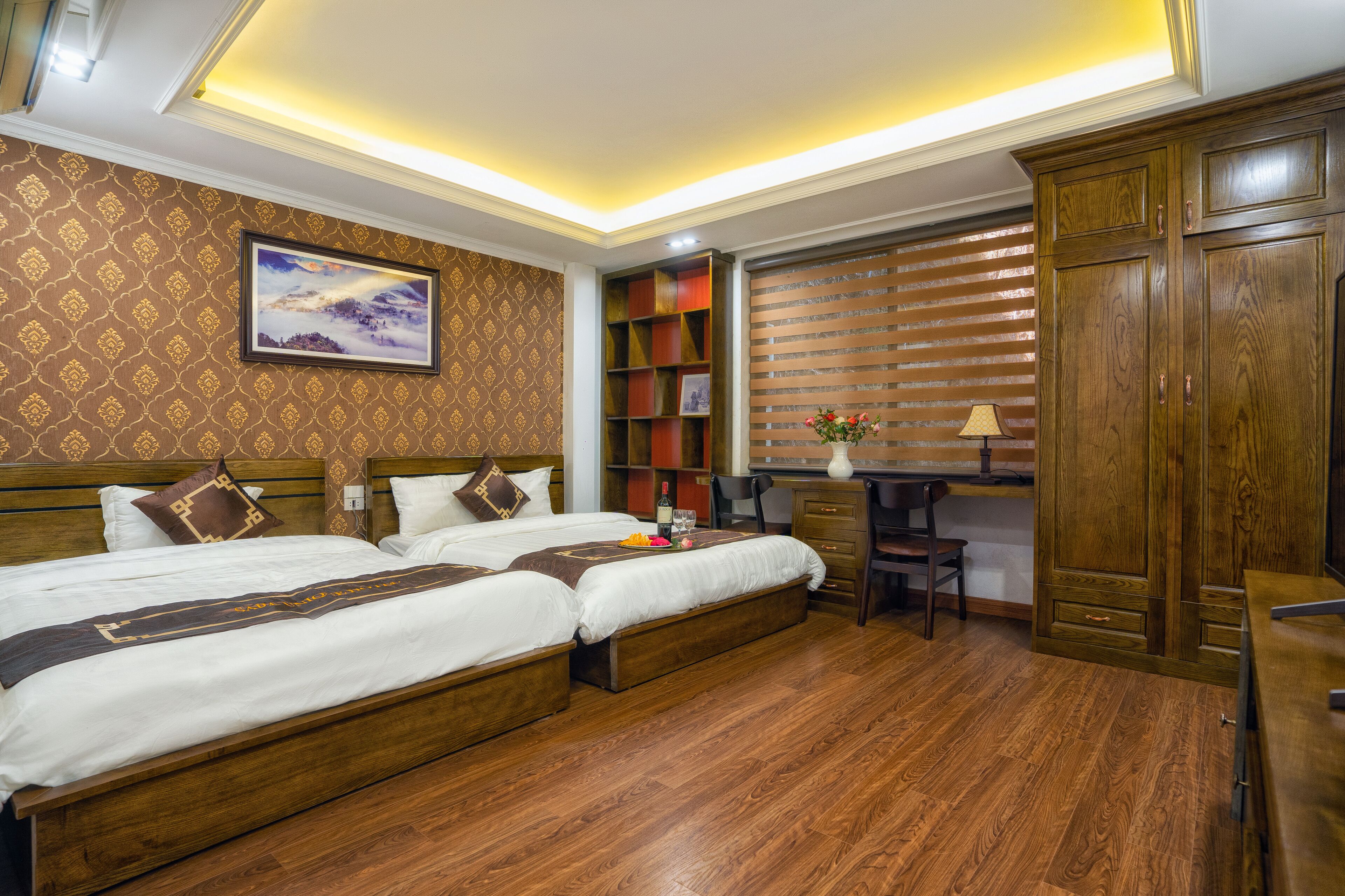A hotel room with two beds, a desk, a chair, a bookshelf, and a large wooden wardrobe.