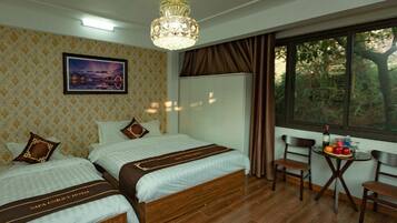 Camera d'albergo con due letti, un lampadario, un quadro, una finestra vista alberi e un tavolino con frutta e una bottiglia.