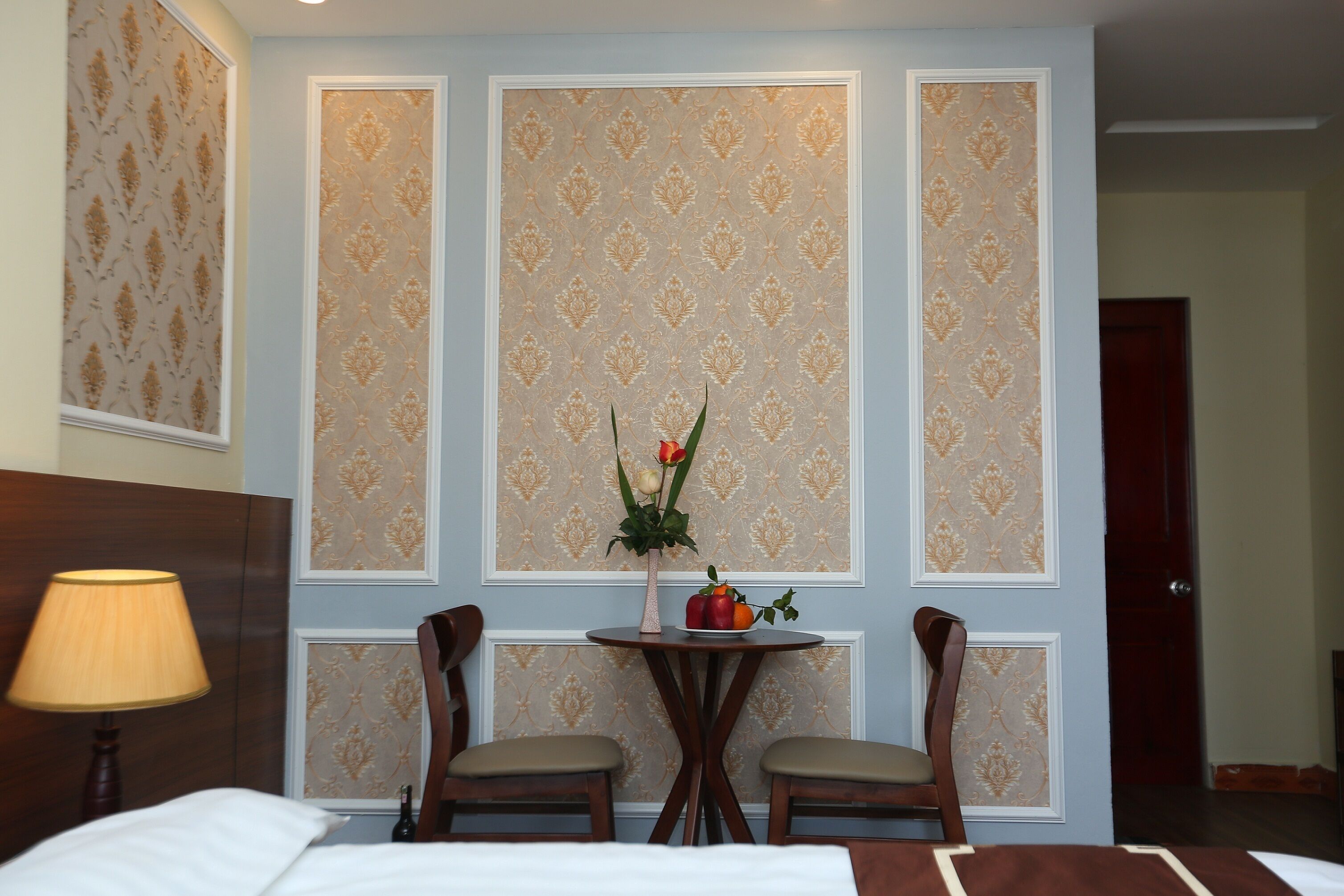 Une chambre d’hôtel avec un lit, deux chaises, une petite table sur laquelle est posé un vase de fleurs et une lampe.