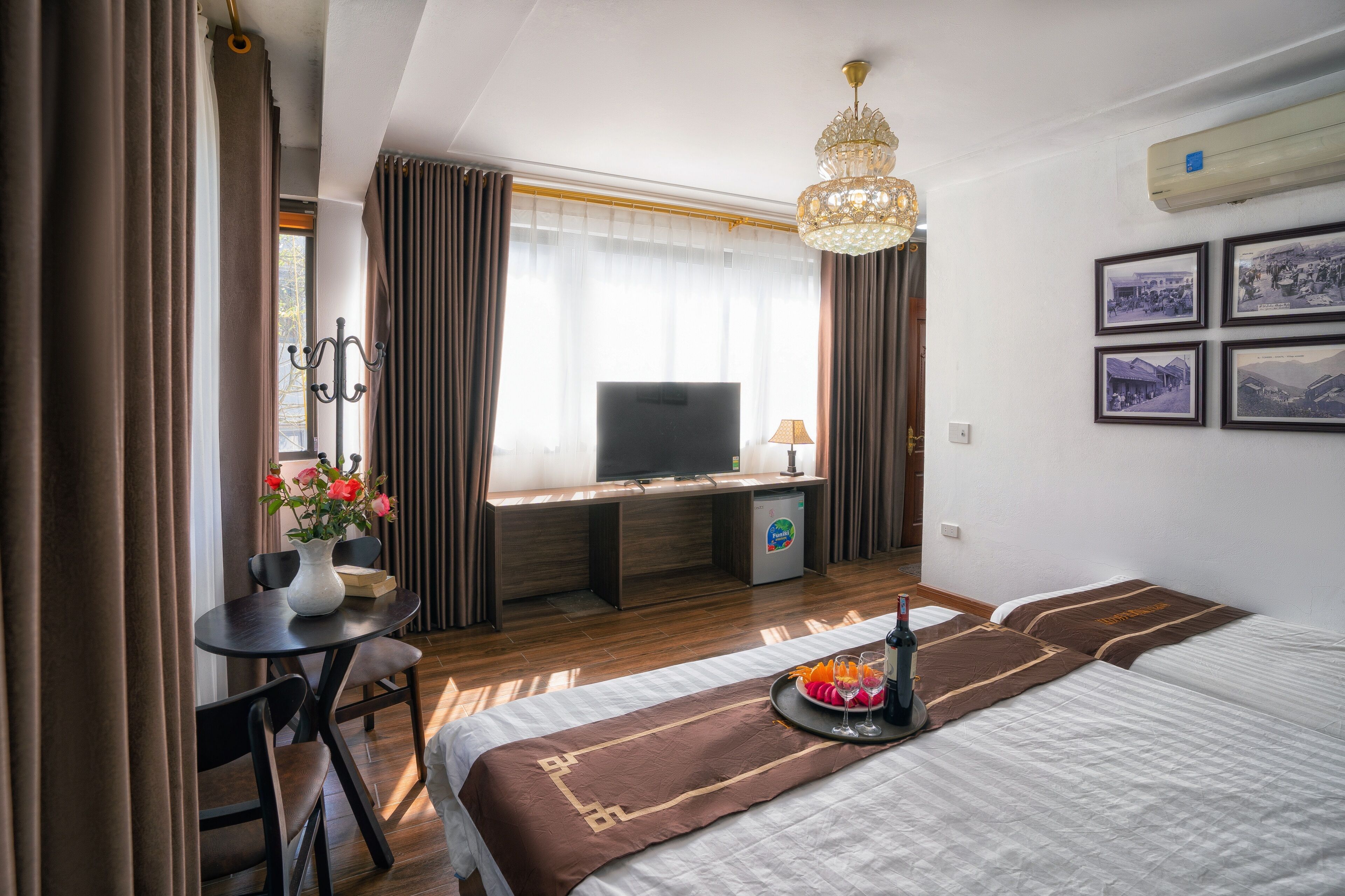 Una camera d'hotel con un letto, una scrivania con televisore, un piccolo tavolino con un vaso di fiori e un minibar.