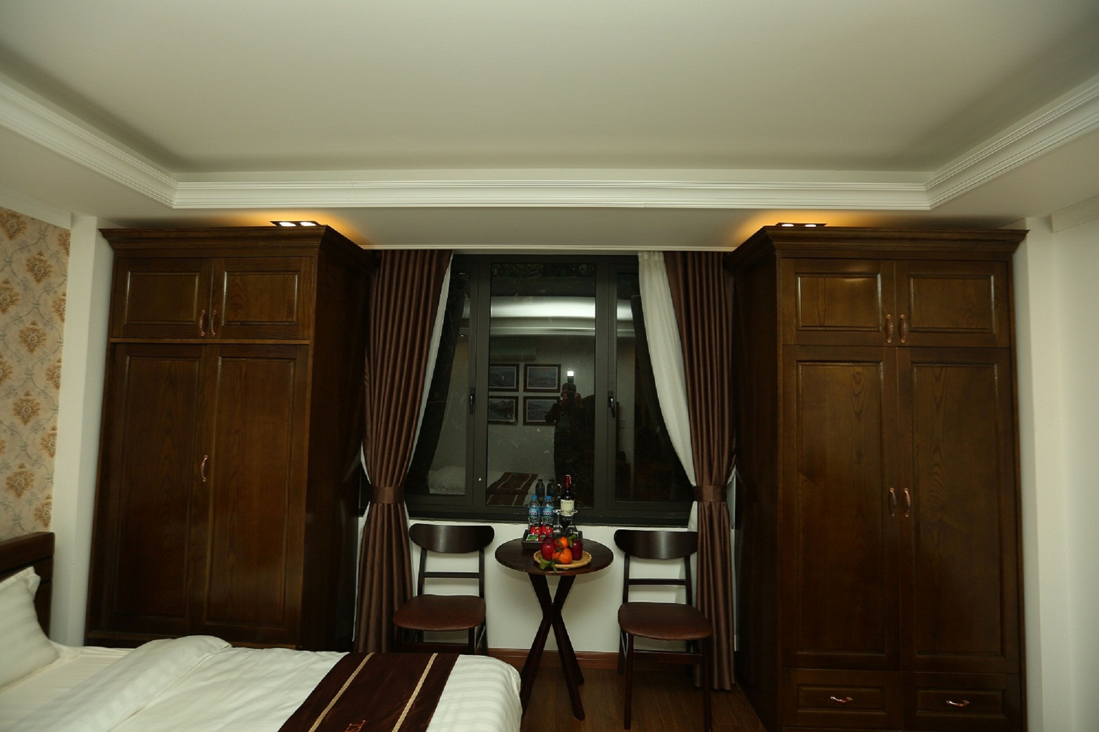 A hotel room with a bed, a small table with fruit, two chairs, and a window with curtains.